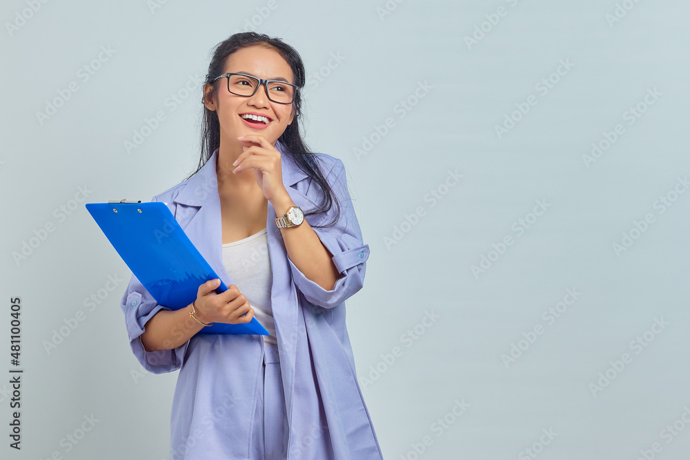 Portrait of cheerful young Asian business woman holding chin while holding document folder isolated on purple background Portrait of cheerful young Asian business woman holding chin while holding document folder isolated on purple background