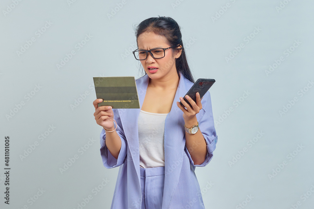 Portrait of surprised young Asian woman looking at vehicle book and holding smartphone isolated on white background Portrait of surprised young Asian woman looking at vehicle book and holding smartphone isolated on white background