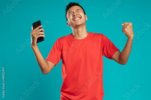 Portrait of a satisfied young man with smartphone celebrating success isolated over blue background