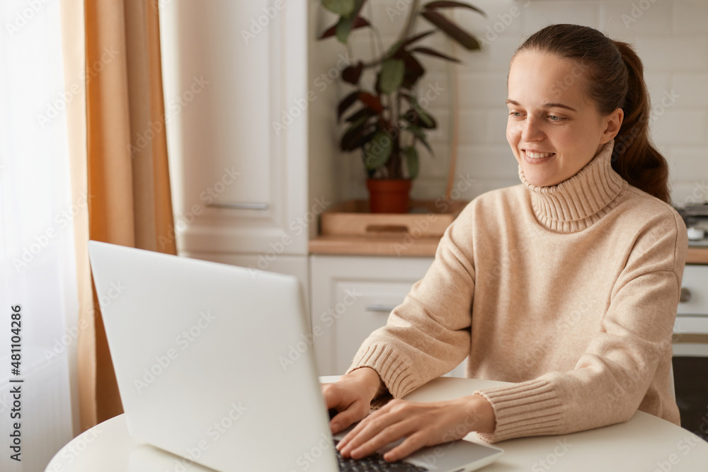 Indoor shot of optimistic happy woman with ponytail hairstyle wearing casual style beige sweater sitting at table in kitchen and working on laptop, typing on keyboard, smiling. Indoor shot of optimistic happy woman with ponytail hairstyle wearing casual style beige sweater sitting at table in kitchen and working on laptop, typing on keyboard, smiling.