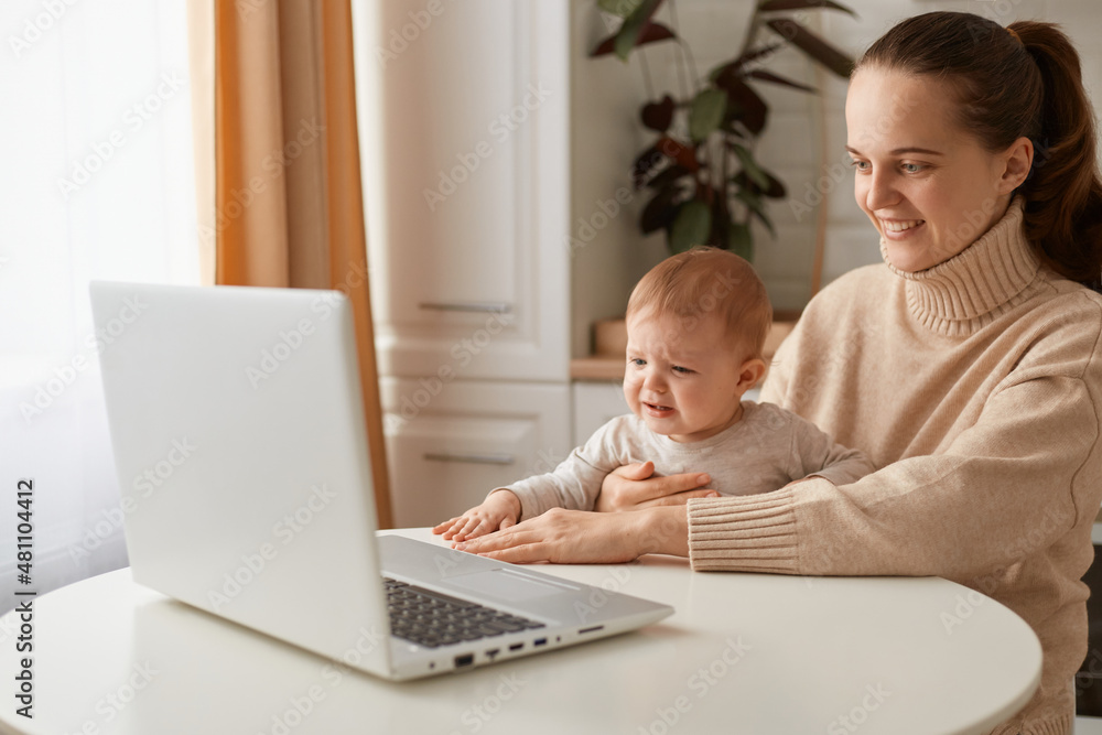 Indoor shot of smiling happy woman with ponytail hairstyle wearing casual style beige sweater sitting at table in kitchen with her toddler baby and looking at laptop display with positive expression. Indoor shot of smiling happy woman with ponytail hairstyle wearing casual style beige sweater sitting at table in kitchen with her toddler baby and looking at laptop display with positive expression.