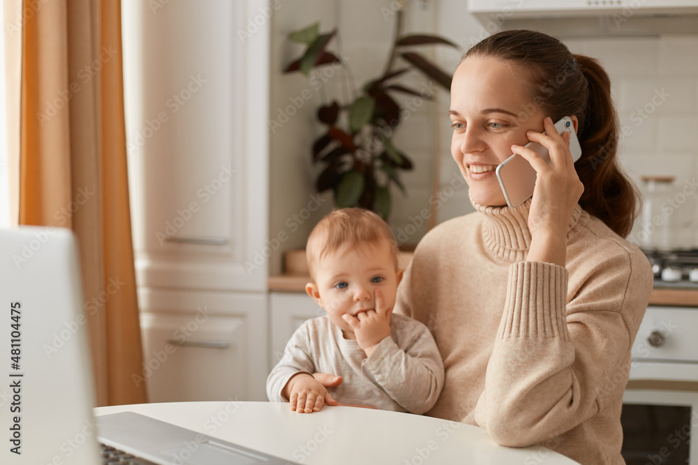 Indoor shot of happy smiling woman wearing beige casual style sweater with ponytail hairstyle sitting at table with her toddler daughter and talking phone with optimistic facial expression. Indoor shot of happy smiling woman wearing beige casual style sweater with ponytail hairstyle sitting at table with her toddler daughter and talking phone with optimistic facial expression.