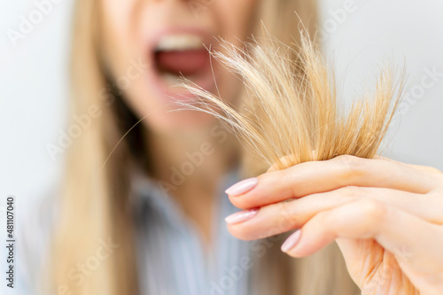 A shocked and upset blond woman holds the damaged brittle dry split ends of her long hair in her hand in front of her face, her mouth wide open in surprise, close-up. Health care and haircare concept. photo
