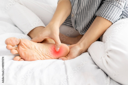 Young woman feeling pain when she press on heel and foot while sitting on the bed at home. Painful female legs from wearing uncomfortable tight shoes and high heels, sprain, arthrosis. Health concept. photo