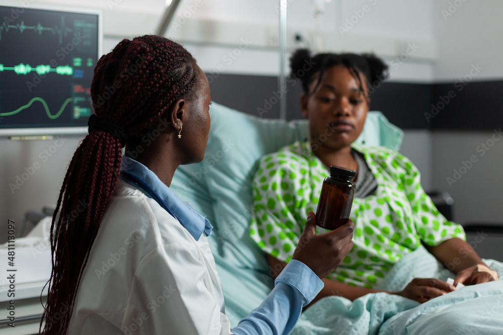 African american practitioner doctor standing beside sick patient holding flu pills bottle explaining healthcare treatment during medical examination in hospital ward. Medicine services African american practitioner doctor standing beside sick patient holding flu pills bottle explaining healthcare treatment during medical examination in hospital ward. Medicine services