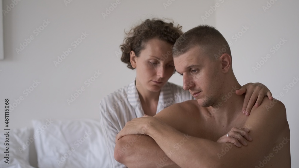 Fototapeta premium Thoughtful depressed young man sitting in bed while his girlfriend comes and hugs him to support, marriage intimate connection