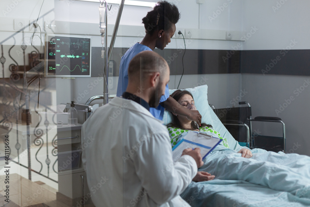 Fototapeta premium Through the window view of nurse putting oxygen mask on unconscious female patient in modern hospital ward. Consulting doctor writing on clipboard with medical lab results while caregiver attends.