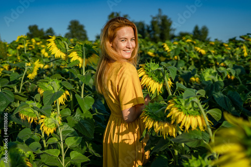 Portrait of a happy young red-haired lady in a yellow dress in a sunflower field on a sunny day. The girl turned towards the camera smiling cheerfully.