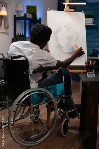 Young artist designer in wheelchair drawing vase on canvas working at sketch making shadows using graphic pencil during art lesson. Illustrator man with disability creating masterpiece