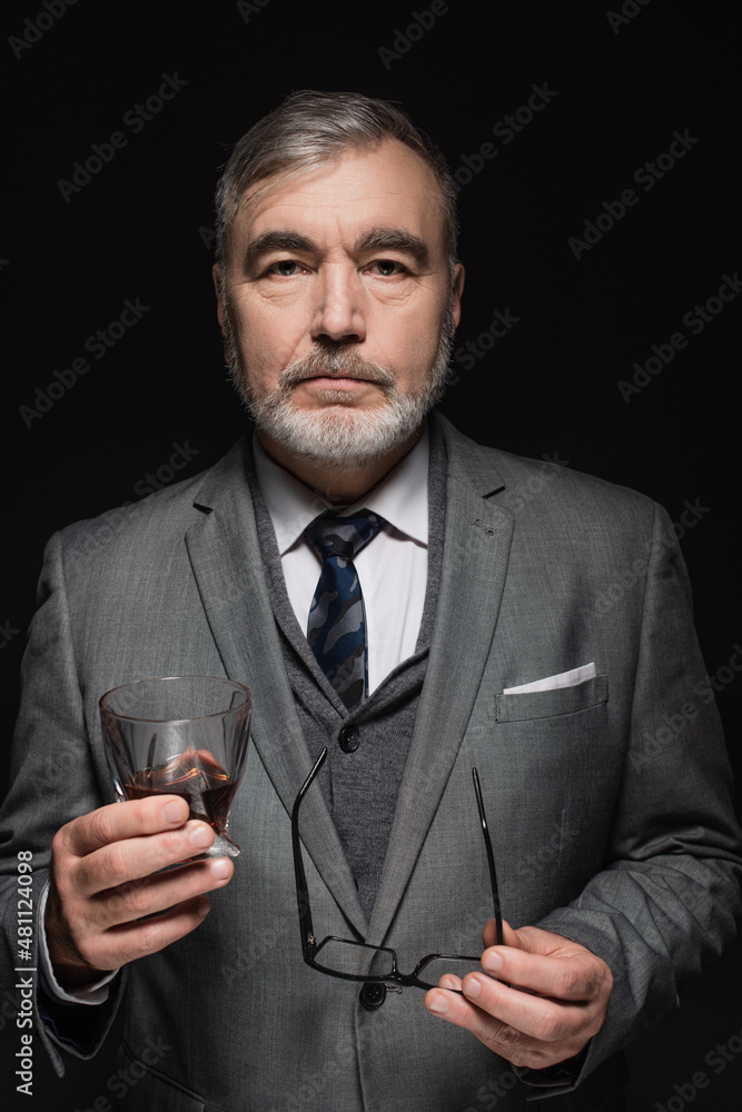 stylish senior man with glass of whiskey and eyeglasses looking at camera isolated on black. stylish senior man with glass of whiskey and eyeglasses looking at camera isolated on black.
