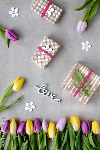 Fototapeta Naklejka Na Ścianę i Meble -  Gifts wrapped in craft paper, tulips in various colors. Yellow, magenta, pink flowers. Geometric Spring flat lay, light grey stone background. Easter, Mothers day, March 8 International Women's day.