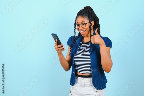 Εκτύπωση καμβά Happy cheerful Afro American woman looks at screen of smart phone enjoys online chatting types text message surfs social networks dressed casually poses against blue background