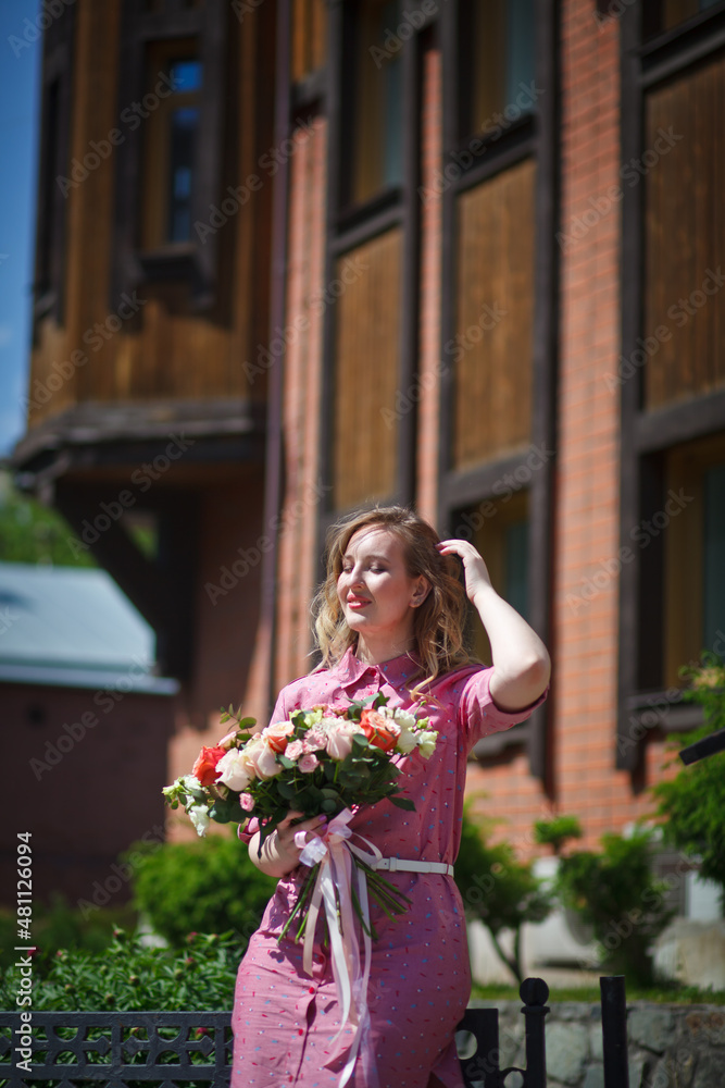 Fototapeta premium Young happy blonde woman in a pink dress with a large bouquet of flowers walks around the old European city and enjoys life on a summer day, beautiful portraits with a huge bouquet of flowers.