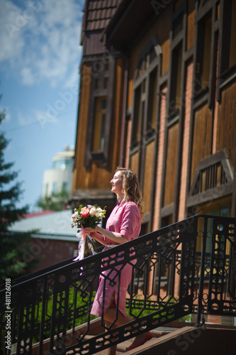 Young happy blonde woman in a pink dress with a large bouquet of flowers walks around the old European city and enjoys life on a summer day, beautiful portraits with a huge bouquet of flowers. © AKaltykova