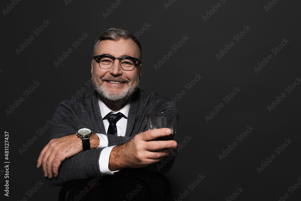 happy senior man in stylish clothes holding glass of whiskey and looking at camera while sitting isolated on dark grey.