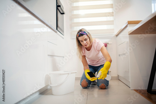 A maid kneeling in kitchen and cleaning floor with detergent.