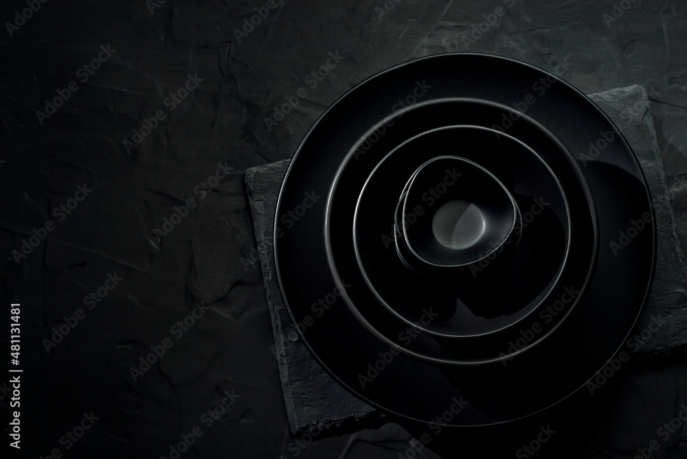 Obraz premium Black tableware on a dark background. Plates and bowls of different shapes and sizes are stacked in a pile. Contrasting dramatic light as an artistic effect.