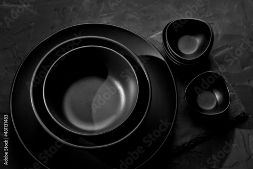 Black tableware on a dark background. Plates and bowls of different shapes and sizes are stacked in a pile. Contrasting dramatic light as an artistic effect.