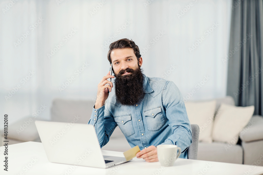 A bearded man sitting at home, having a phone call and preparing to use his platinum credit card. A bearded man sitting at home, having a phone call and preparing to use his platinum credit card.