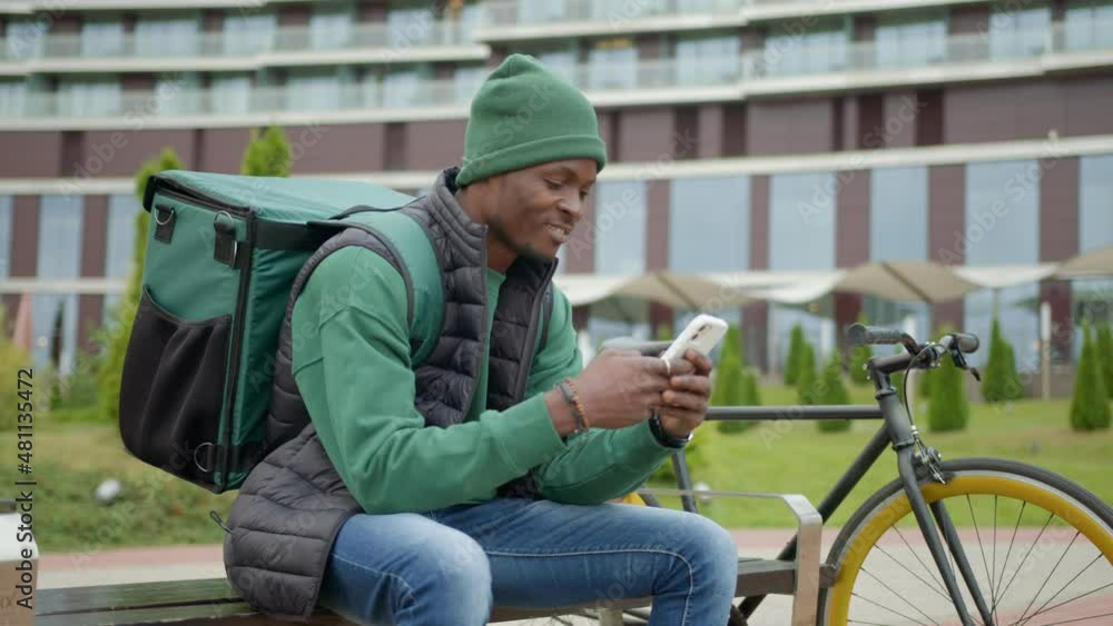 A young black delivery man in a city park on a bench takes an order on his phone. A young black delivery man in a city park on a bench takes an order on his phone.