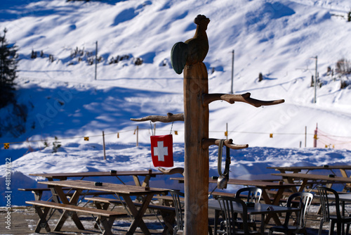 Painted Swiss flag on wood with wooden pole and bell and wooden rooster at terrace of restaurant at Mürren Winteregg on a sunny winter day. Photo taken January 15th, 2022, Lauterbrunnen, Switzerland. photo