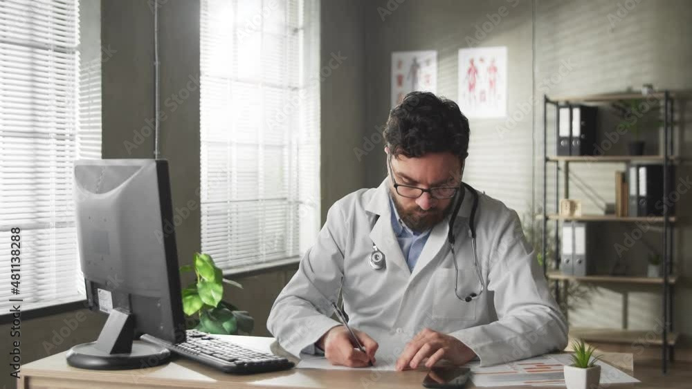 A young doctor works at a computer in a hospital office and writes down data on paper. Telemedicine concept A young doctor works at a computer in a hospital office and writes down data on paper. Telemedicine concept