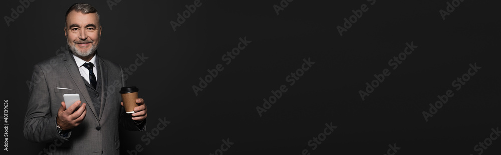 happy senior businessman holding mobile phone and paper cup while looking at camera isolated on black, banner. happy senior businessman holding mobile phone and paper cup while looking at camera isolated on black, banner.