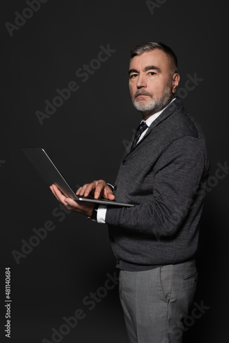senior bearded man in jumper holding laptop and looking at camera on dark grey.