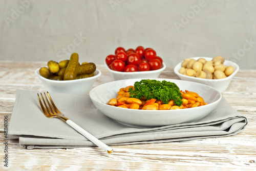Beans with carrots in a white bowl. Beans with pickled tomatoes, mushrooms and cucumbers on a white background. Food on a shabby table. Bowl on an old wooden board.