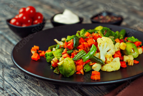 Mix of vegetables close up. Salad of various chopped vegetables. Roasted cabbage, carrots, broccoli, peas in a black plate. Food on a wooden board. Marinated tomatoes in the background.