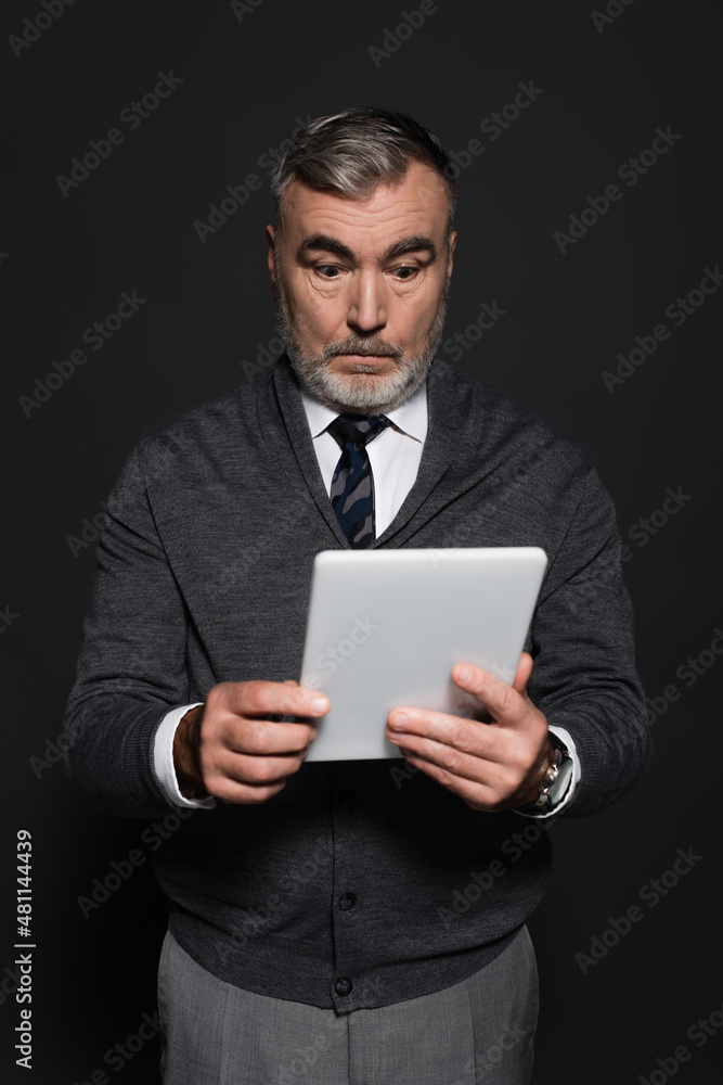 surprised senior man in jumper and tie looking at digital tablet isolated on dark grey. surprised senior man in jumper and tie looking at digital tablet isolated on dark grey.