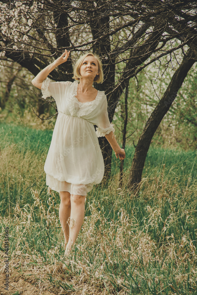 Beautiful young woman in a white chiffon dress with a short blonde haircut looks at a blooming cherry branch Beautiful young woman in a white chiffon dress with a short blonde haircut looks at a blooming cherry branch