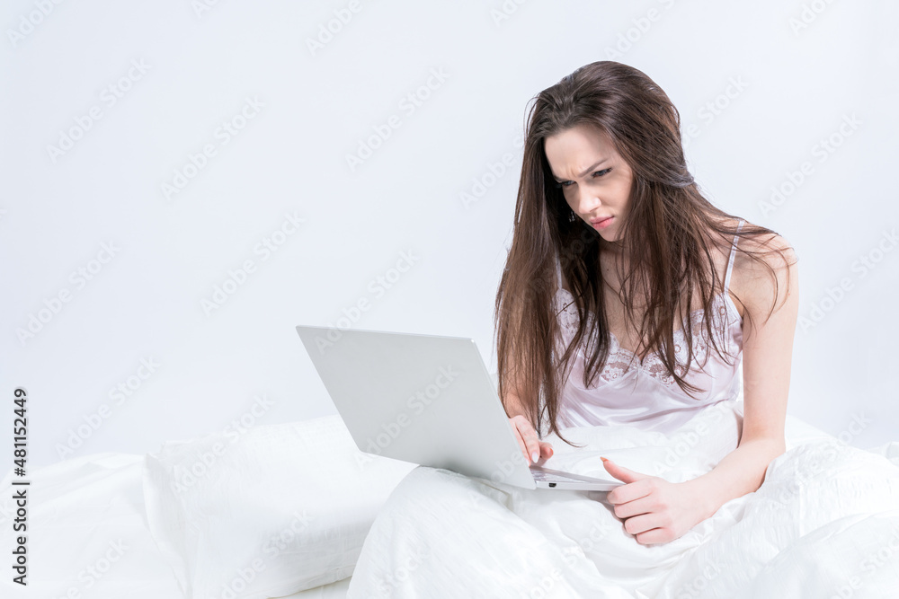 Good morning. Conversation via video link. A brunette girl sits on a white bed in her pajamas, holds a laptop in her hands. Remote. Woman working at home Good morning. Conversation via video link. A brunette girl sits on a white bed in her pajamas, holds a laptop in her hands. Remote. Woman working at home