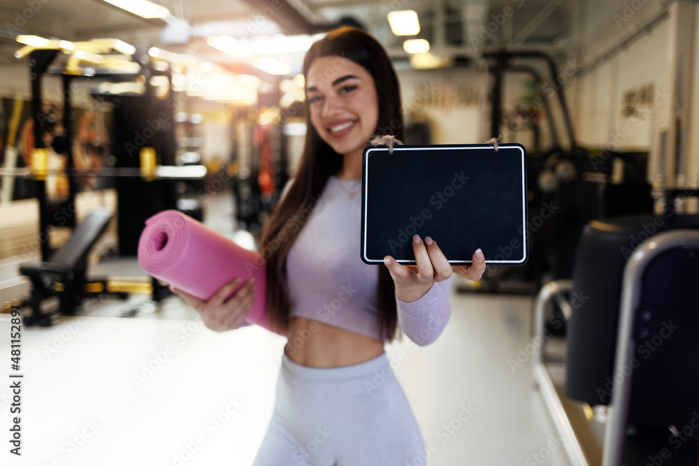 Portrait shot of pretty young Caucasian woman holding a small wooden sign, standing in the gym. Cropped shot of an attractive young woman holding a board in her hands as she enters the gym.