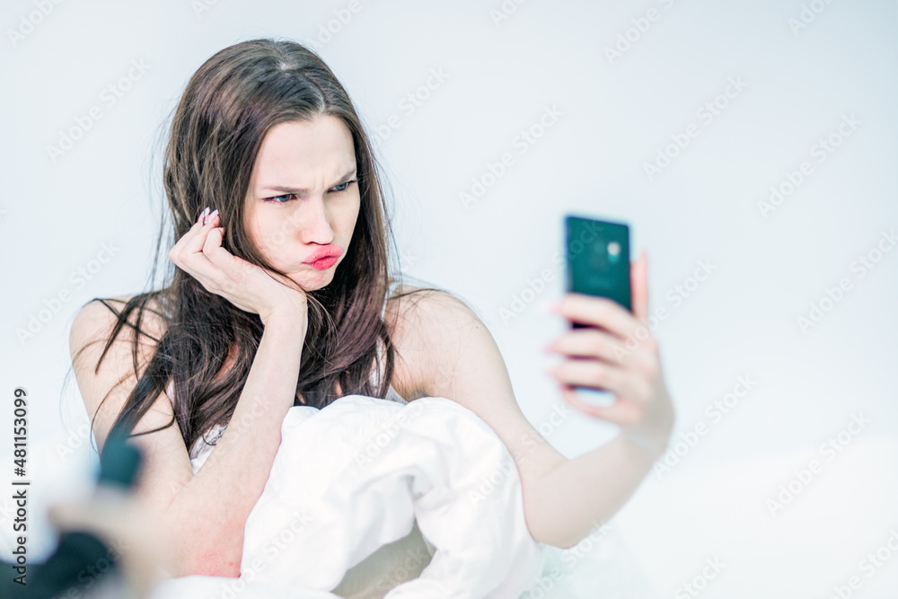 A brunette in her room on the bed with a smartphone in her hands among the white bed linen. The girl makes grimaces into the phone camera, takes a selfie