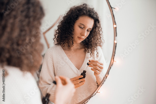 Young woman doing self-care routine at mirror, using aroma oil or serum for skin photo