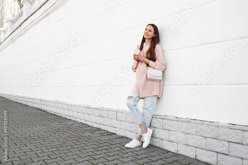 Beautiful young pregnant woman wearing pink sweater, jeans and a bag standing near white wall on a city street with a drink in her hands. © NesolenayaAleksandra