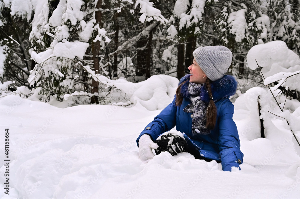 cheerful pretty girl is sitting on her knees in snowdrift and laughing. Outdoor activities in winter forest.
