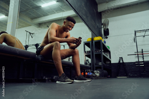 Young African American male looking at his phone to reply to an online message. Happy, mixed race male smiling at his phone while sitting down in the gym. High quality photo 