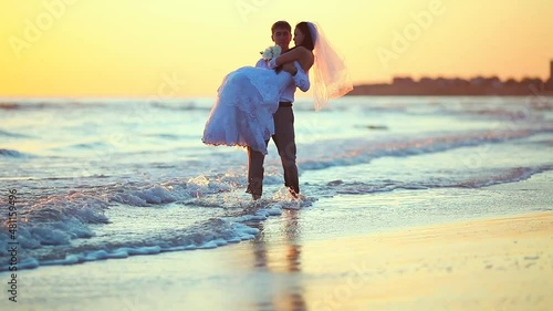 Newly married walking on the sea in the rays of sunset. The groom holds the bride in his arms, kissing and carrying her along the surf line on the sandy beach of the sea on their wedding day.