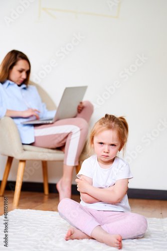 Upset little girl sitting lonesome while mother using laptop. Mother don't have time for daughter