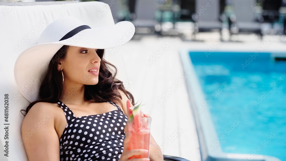 brunette young woman in swimsuit and straw hat holding cocktail while lying on lounger