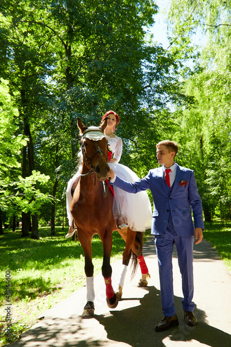 Happy bride and groom with brown horse in green summer sunny park. Weddina walk with big horse