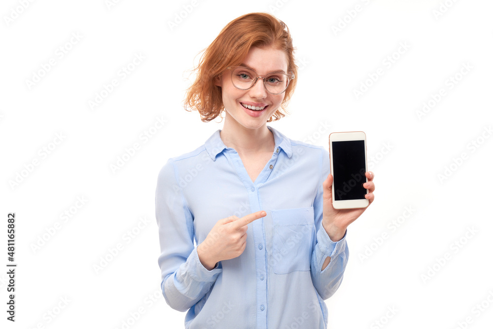 Shocked joyful redhead girl points finger at smartphone blank screen in glasses and business shirt isolated on white studio background Shocked joyful redhead girl points finger at smartphone blank screen in glasses and business shirt isolated on white studio background