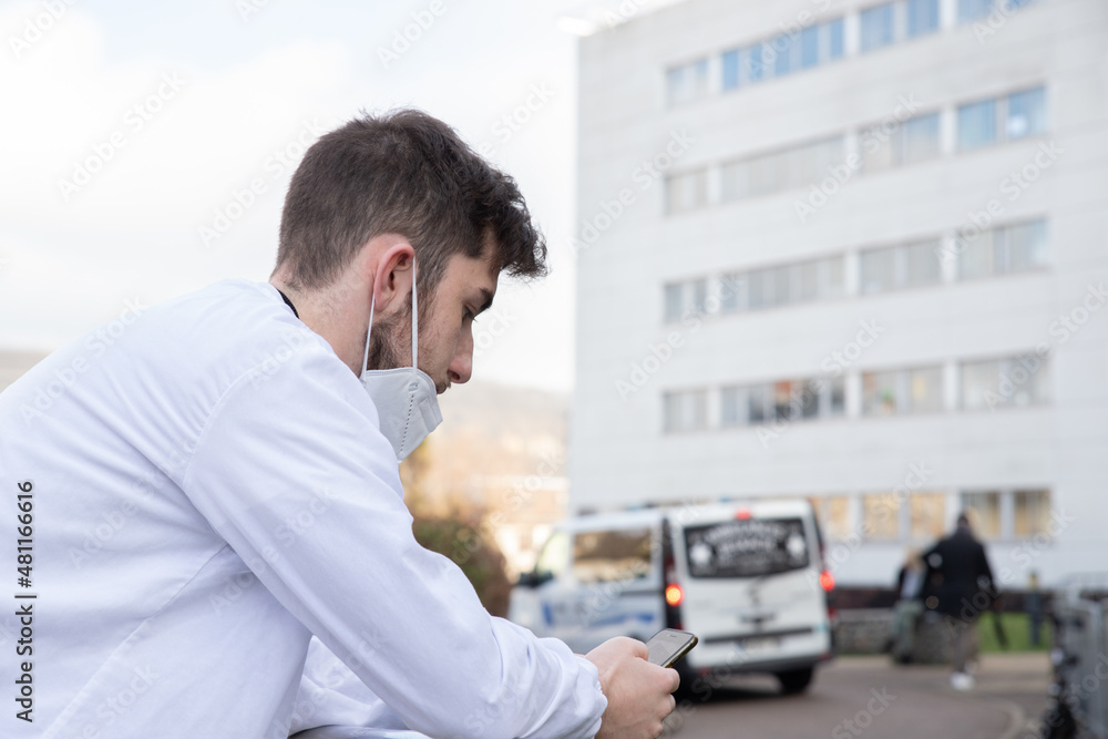 Fototapeta premium Portrait d'un soignant, personnel médical ou infirmier devant un hôpital portant un masque de protection contre le virus Covid 19. Il est fatigué et déprimé. Il regarde son téléphone pendant sa pause