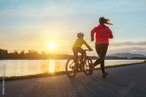 Happy mother and son go in sports outdoors. Boy rides bike in helmets, mom runs on sunny day. Silhouette family at sunset. Fresh air. Health care, authenticity, sense of balance and calmness