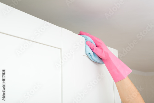 Young adult woman hand in pink rubber protective glove using blue dry rag and wiping dust from top of white wooden wardrobe in room. Closeup. Regular cleanup at home.