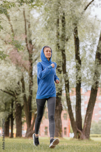 a European woman in a blue hoodie is running and doing sports in the park. fitness and yoga outdoor sports, exercise and meditation and fitness for body health. spiritual development. outdoor sports © MyJuly