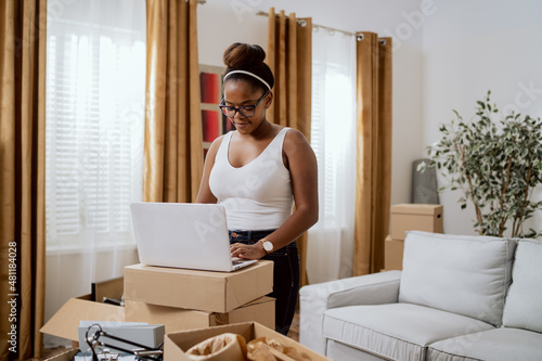 Satisfied woman after moving to new apartment, girlfriend uses laptop that sits on top of cardboard boxes due to lack of furniture, online search for furnishings, online shopping