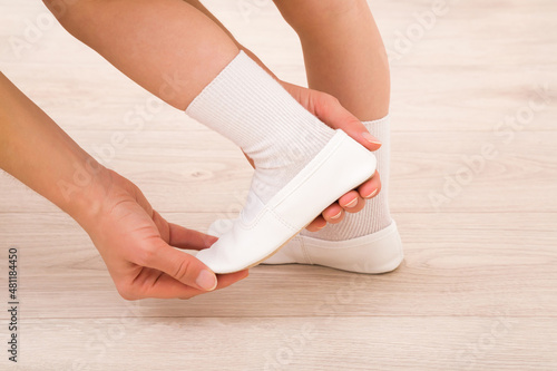 Young adult mother hands putting white leather gymnastic slippers on daughter foot on light wooden floor. Closeup. Side view. Child shoes for sport lesson in kindergarten.